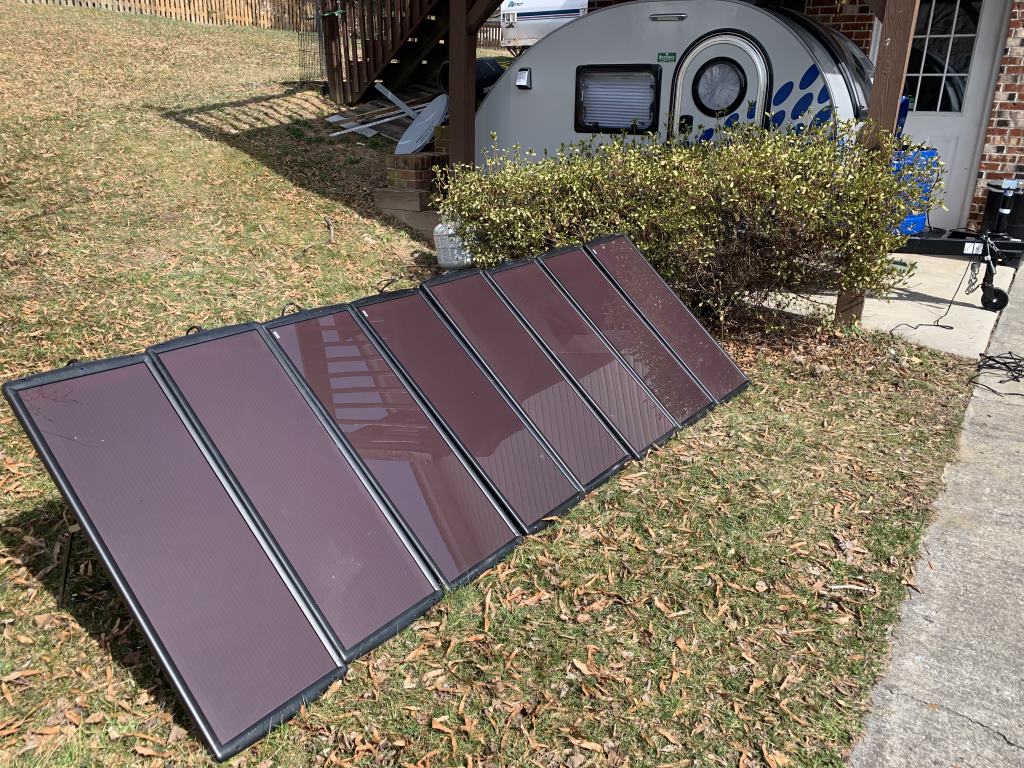 An array of eight solar panels are collecting sunlight in front of a small camping trailer.